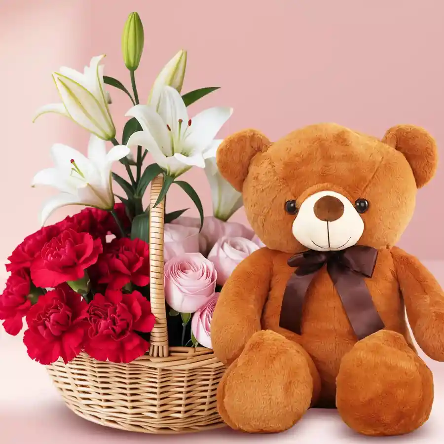 Expressive mixed floral basket featuring red carnations, pink roses, white lilies, and a cute brown teddy bear.