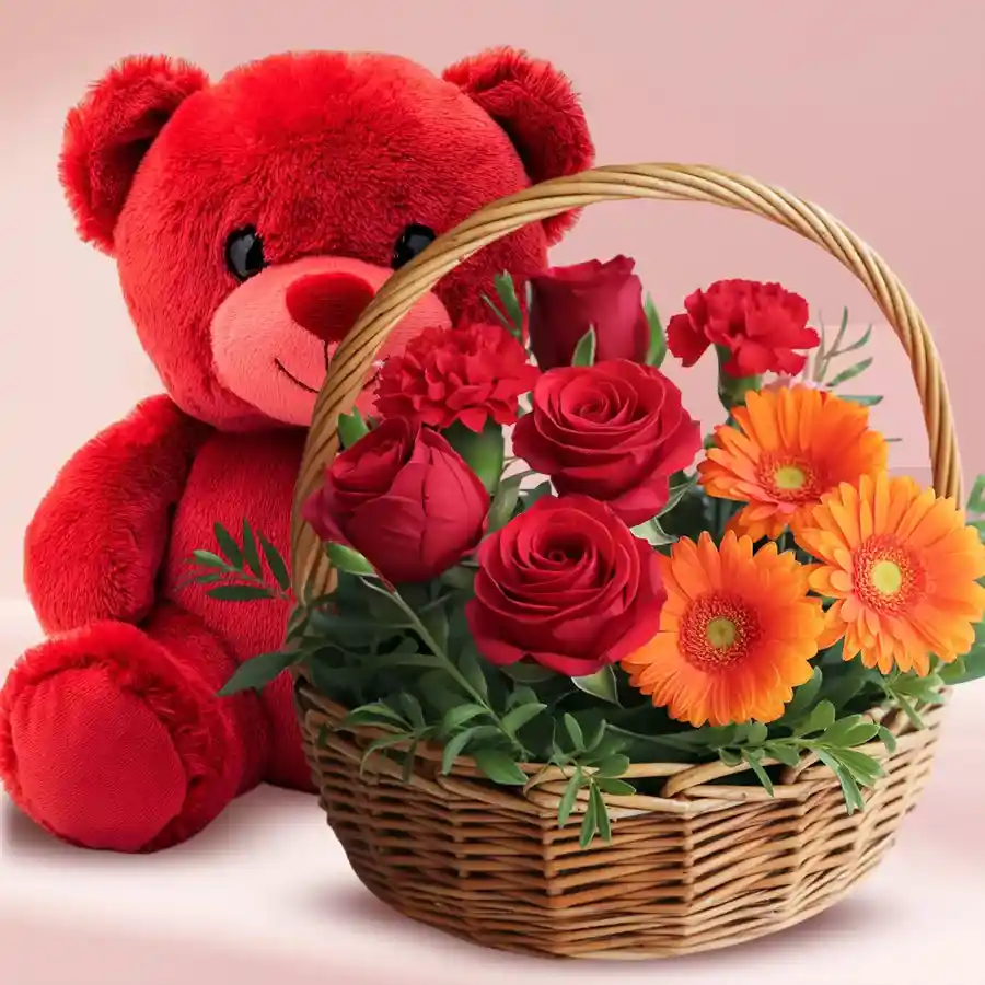 A vibrant floral basket featuring red roses, orange gerberas, pink and red carnations, alongside a cute red teddy bear with a bowtie.