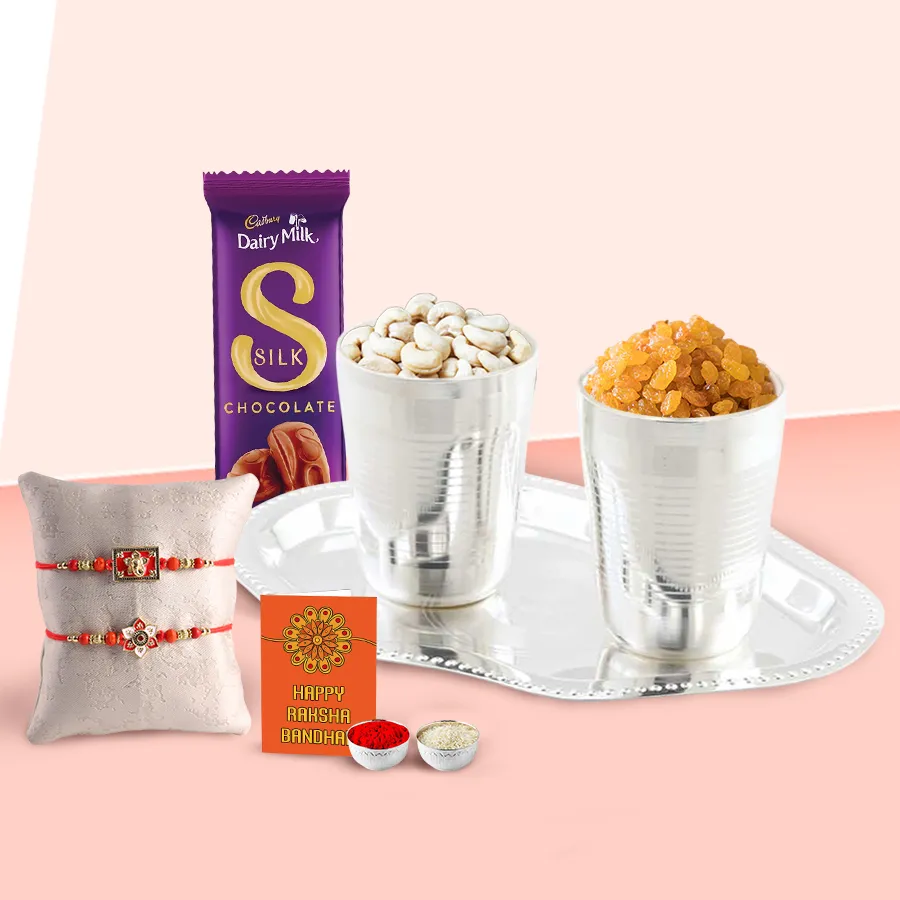 Delicious Dry Fruits Hamper featuring silver-plated glasses and tray, Cadbury Dairy Milk Silk n Family Set, two designer rakhis, and a heartfelt message card with Mouli Rakhi and roli tika chawal.