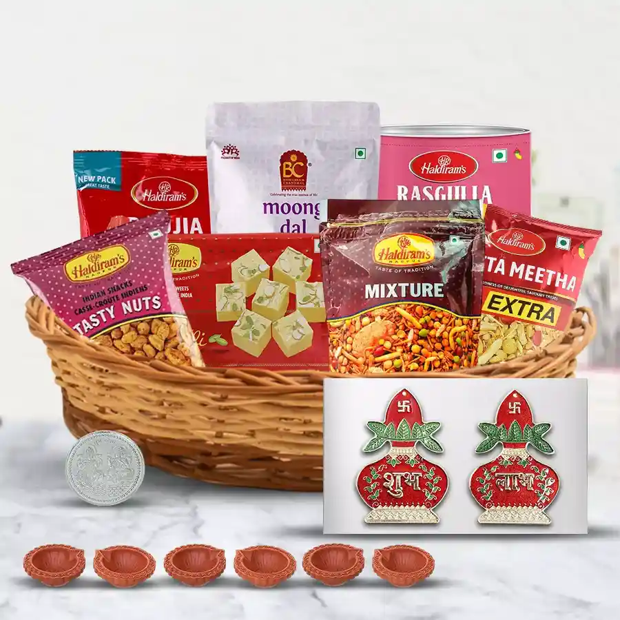 Diwali sweets and snacks hamper from Haldiram featuring a cane basket filled with Badam Pakodi, Khatta Meetha, Soan Papdi, Rasgulla, and Bhujia, accompanied by decorated diyas, Subh Labh wall stickers, a decorative diya set, and a silver-plated Ganesh-Laxmi coin.