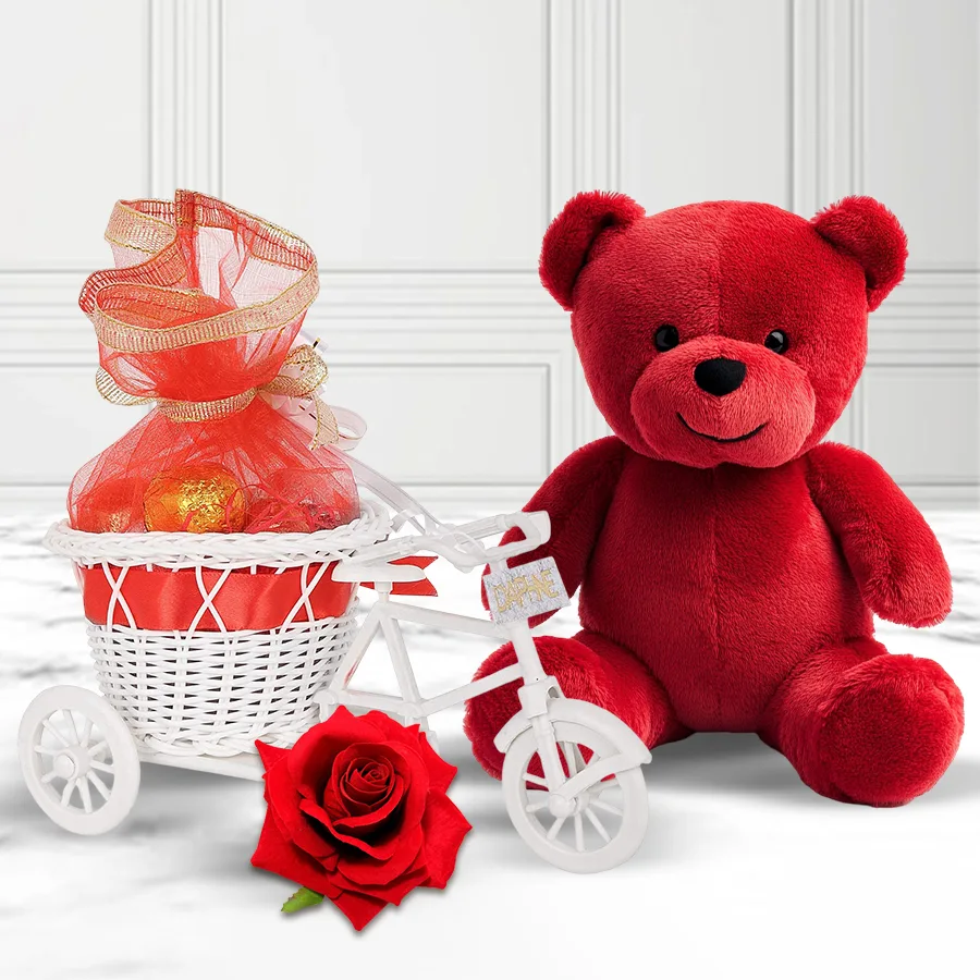 Charming red teddy in a tri-cycle surrounded by heart-shaped chocolates and a red velvet rose