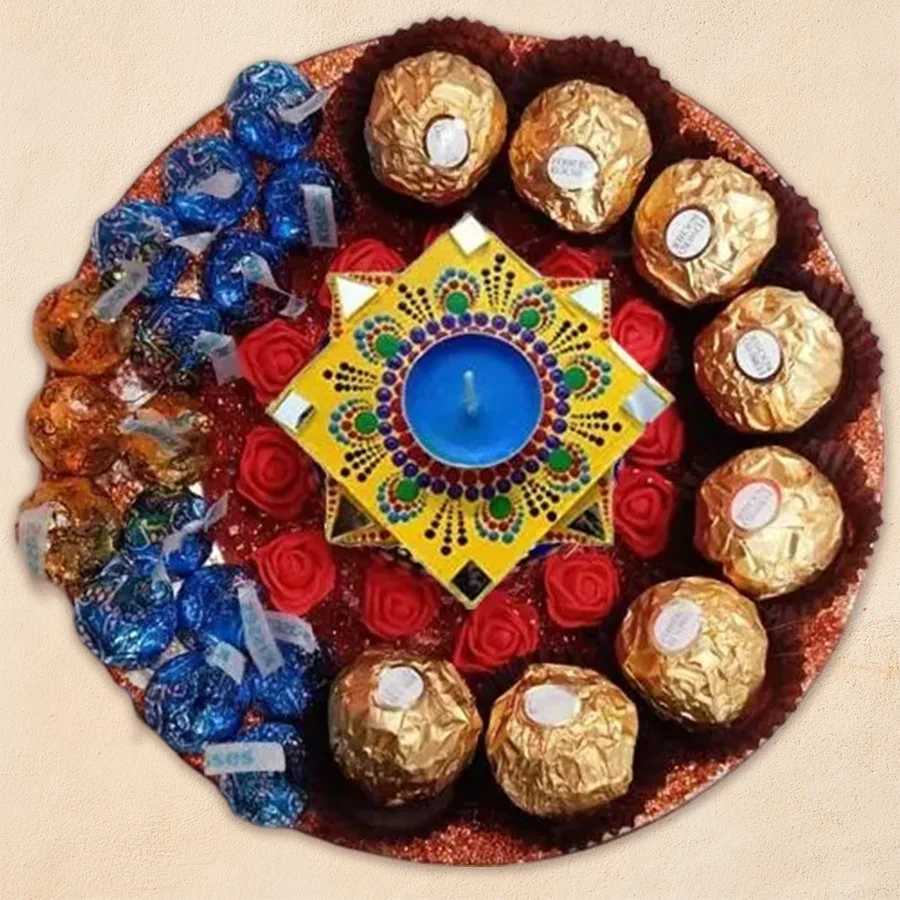 A beautifully arranged platter featuring imported chocolates, including Ferrero Rocher and Hershey's Kisses, alongside a handcrafted mandala-decorated diya on an elegant steel thali, adorned with artful flowers.