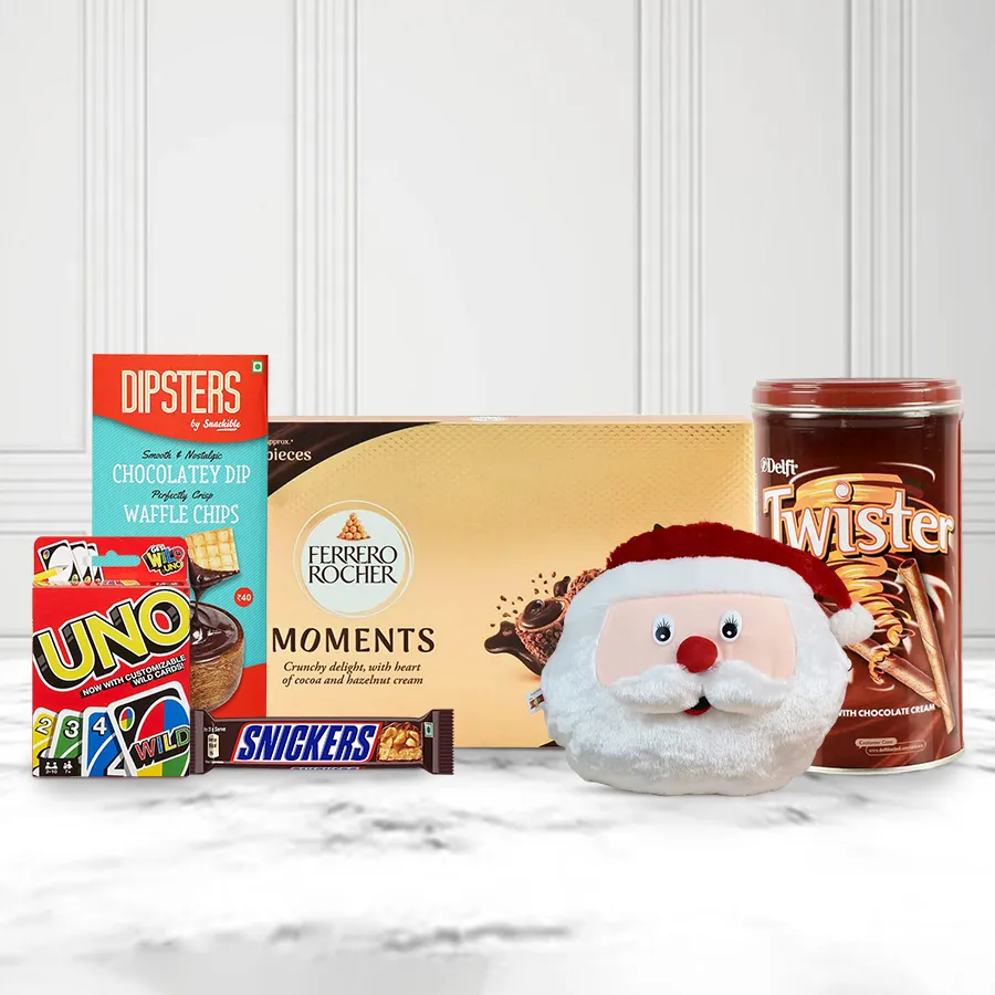 A colorful gift pack featuring a Santa Claus face cushion, assorted chocolates including Ferrero Moments, Delfi Wafers, a Snickers bar, Snackible Dipsters with waffle chips, and a Mattel Uno card game, all arranged beautifully for the holiday season.