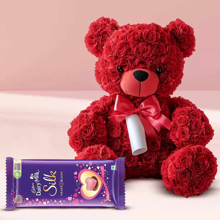 Elegant teddy bear adorned with roses in red, blue, white, or pink, accompanied by a personalized message scroll and Cadbury Silk Heart Blush Chocolate.
