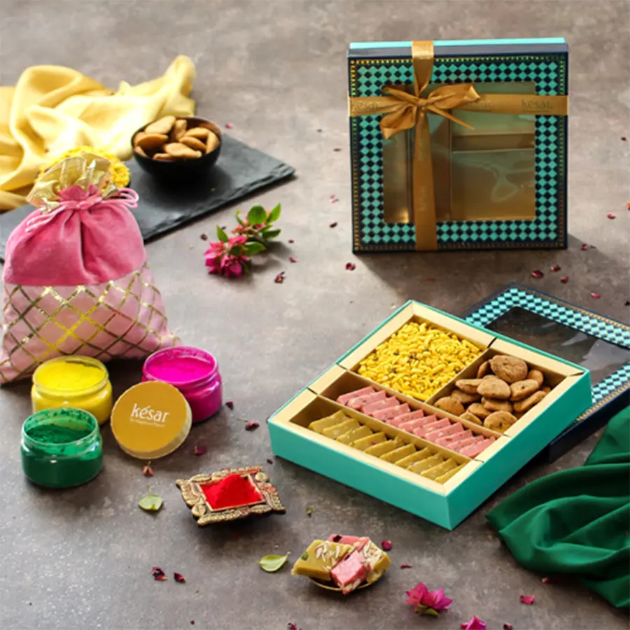 Luscious Sweet N Savories with Herbal Color Combo gift box featuring Thandai Katli, Rose Katli, Methi Suali, Navratan Mixture, herbal colors in a potli, and a Holi greeting card, all presented in a blue window box.