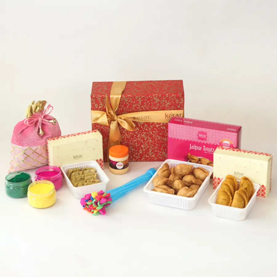 Holi celebration hamper featuring crispy mini samosas, sweet kesar gujiyas, thandai katli, vibrant herbal colors, a playful water balloon, a special balti and pichkari, chandan incense, and a Holi greetings card, all in a handmade paper box.