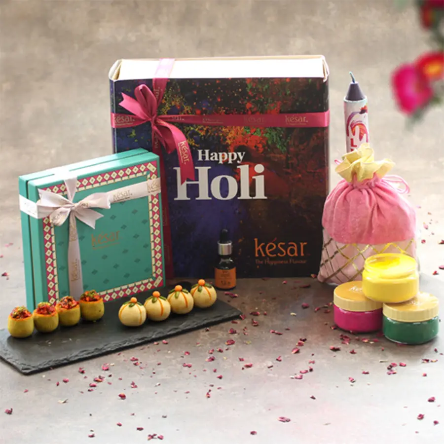 Gift hamper featuring colorful ladoos, herbal powders, a pichkari, essential oil, a color bomb, and a greeting card in a vibrant box for Holi celebration.