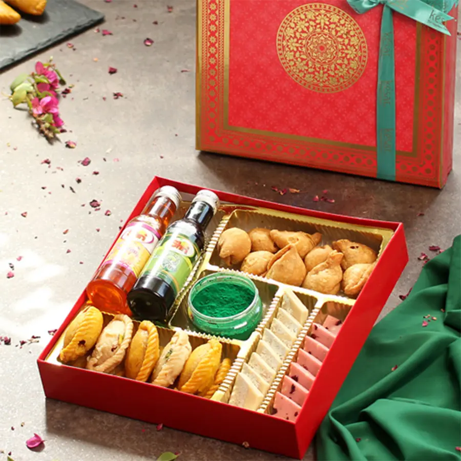 A colorful gift hamper filled with assorted Holi sweets including gujiyas, katlis, mini samosas, two bottles of sharbat, a gulab jar, and a greeting card, all presented in a festive box.