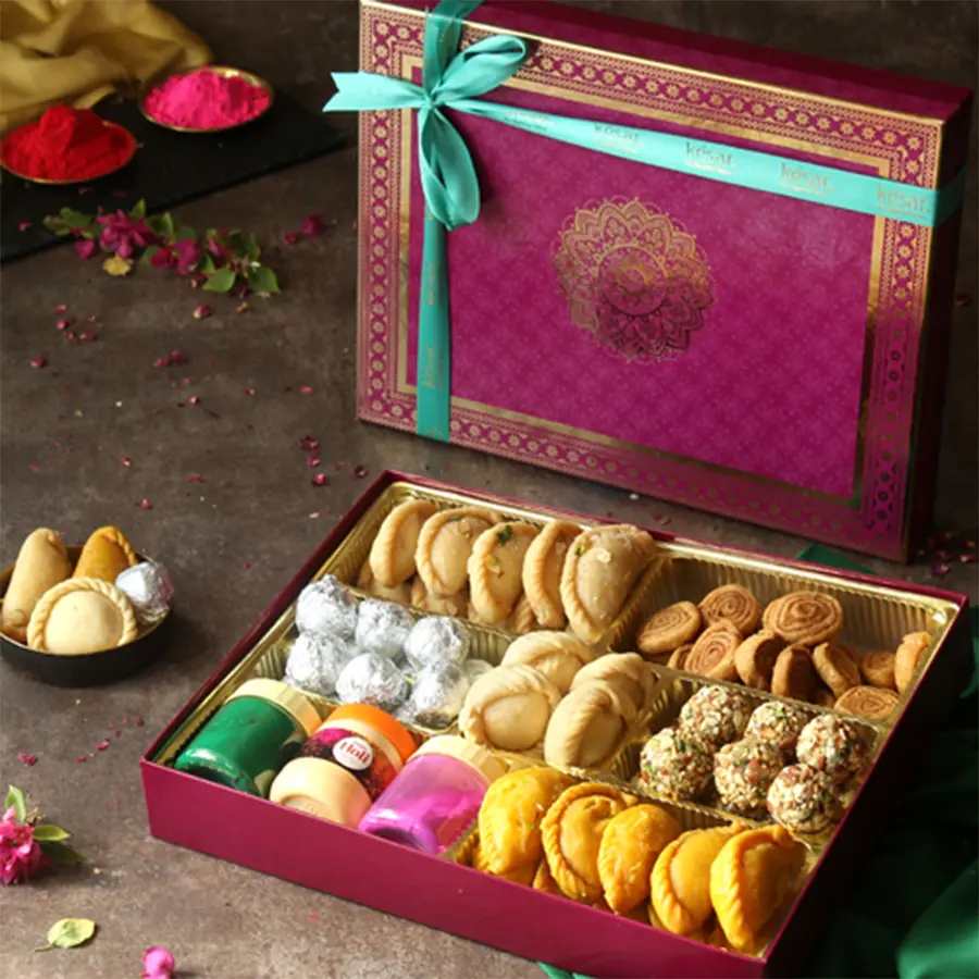 Gift box filled with Holi treats including Chandrakala, Gujiya, Ladoo, Kachori, Bhakarwadi, colorful Gulal, and a Holi greetings card.