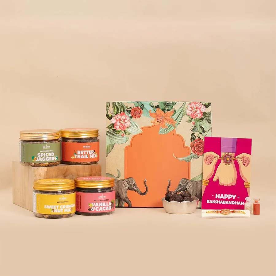 Glam Rakhi Hamper featuring a handcrafted Rakhi, vegan dry fruits, chocolate and vanilla laddoos, nut mixes, spiced jaggery seeds, elegant glass bottles with Roli, Tilak, and Chawal, and a heartfelt greeting card, all beautifully arranged in a modern presentation.