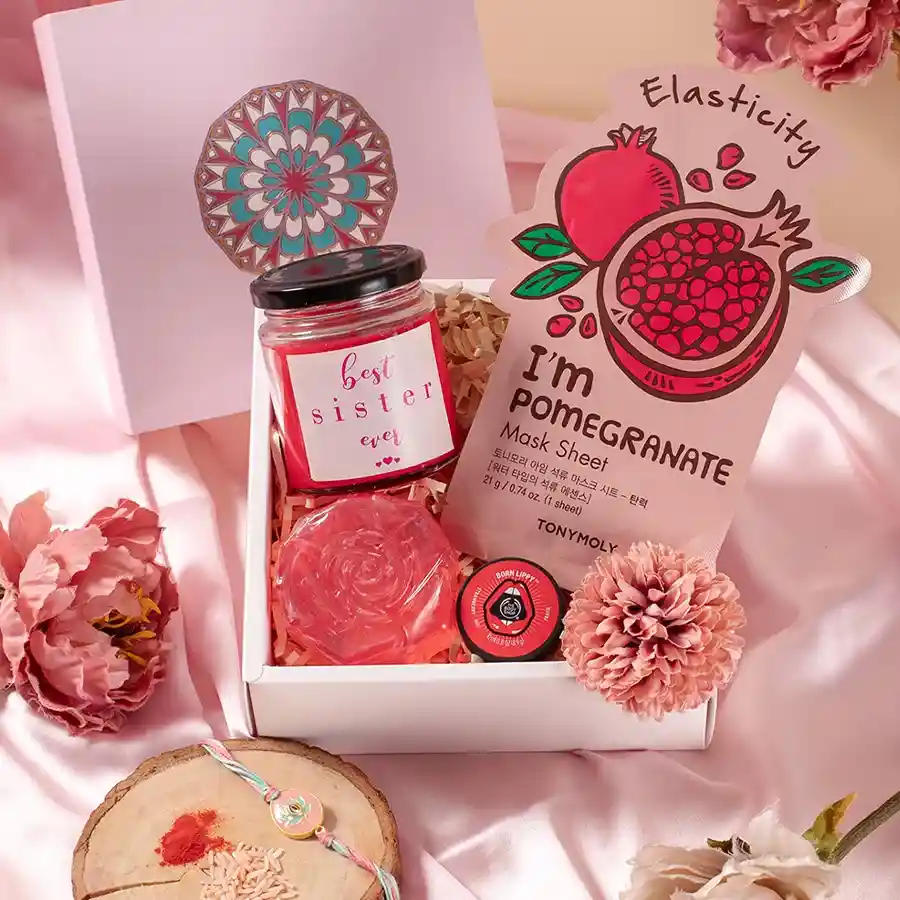 Marvellous Pink Confetti Box of Rakhi Goodies with Pomegranate Mask Sheets, Rose Scented Soap, Rose Candle, Strawberry Lip Balm, and Fancy Rakhi arranged inside a reusable box