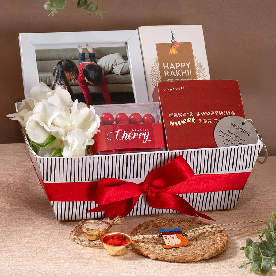 Cherry Dragee Rakhi Hamper featuring a personalized white photo frame, Happy Rakhi chocolate bar, cherry dragees, handmade cookies, a brother keychain, a reusable Kappy tray, a flower arrangement, and a king rakhi.