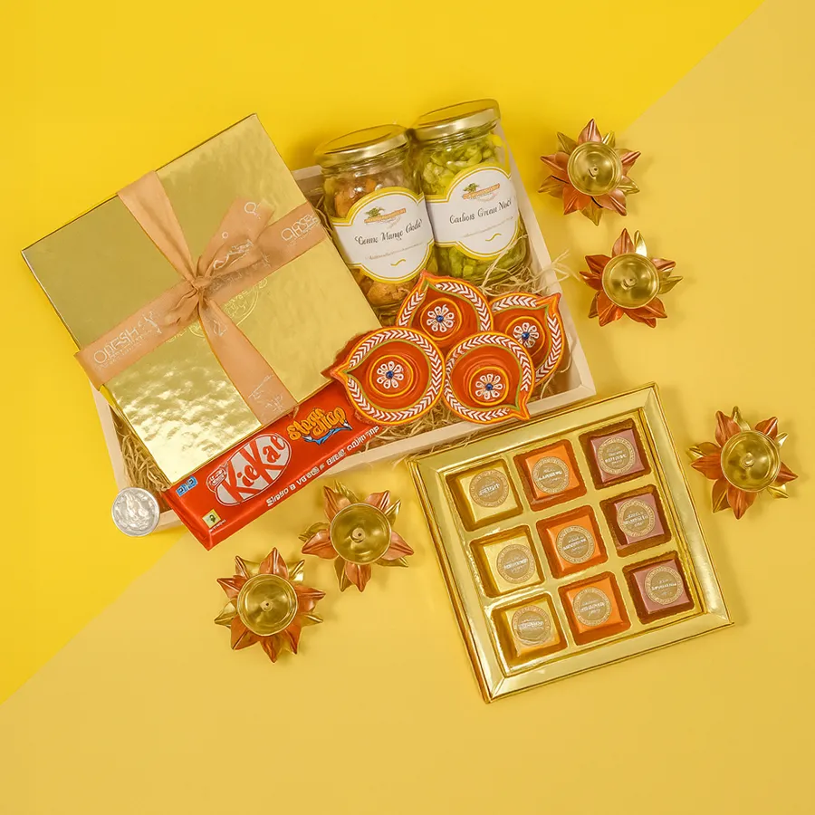 A Taste of Diwali Magic Gift Box featuring cashew green chutney, spicy kismis orange chilies, assorted fudges, a KitKat chocolate, brass and clay diyas, a silver Ganesh Lakshmi coin, and a wooden tray.