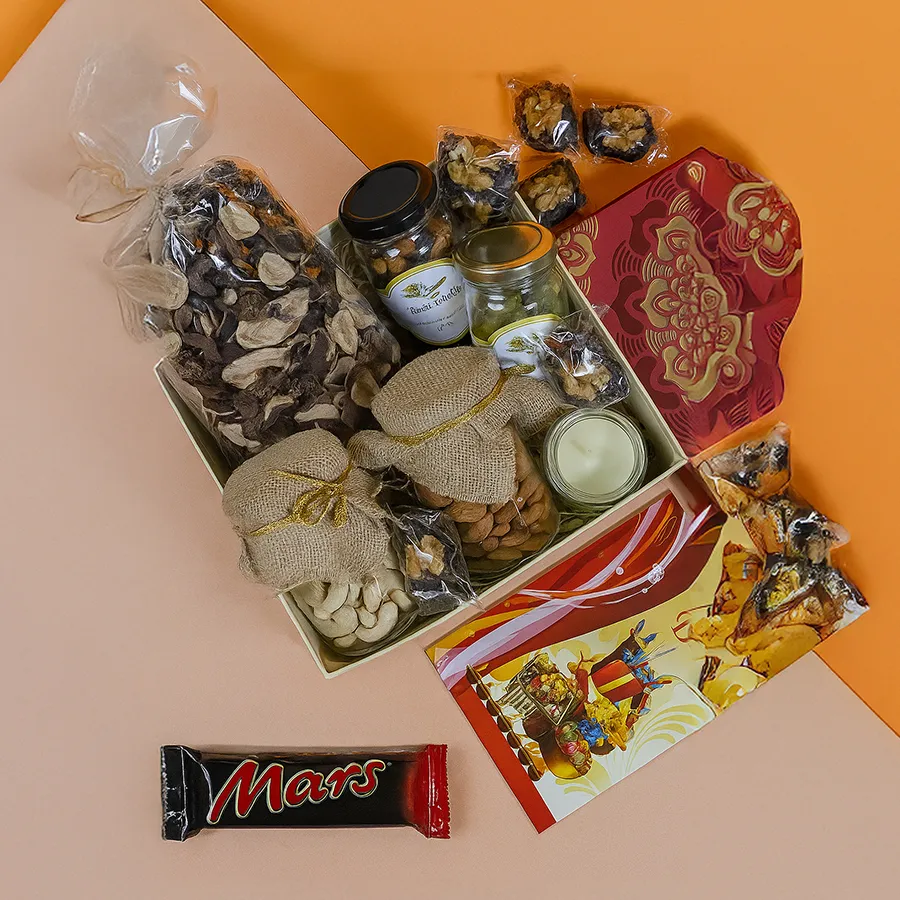 A beautifully arranged Diwali hamper featuring elegant glass jars filled with cashews, almonds, exotic trail mix, walnut-stuffed dates, musk-scented potpourri, and a Bella Vita organic candle, all presented in a reusable Mandala box with a Happy Diwali card.