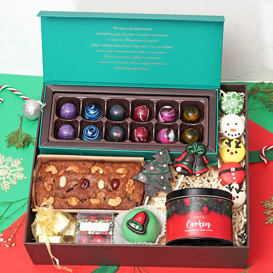 A festive assortment of holiday treats including Bon Bon Chocolates, Christmas Tree and Bell lollipops, a Fruit & Nut Tea Cake, colorful macarons, snickerdoodle cookies in a tin, a Hot Chocolate Bomb, cranberry dragees, and assorted chocolates, all beautifully arranged in a gift box.