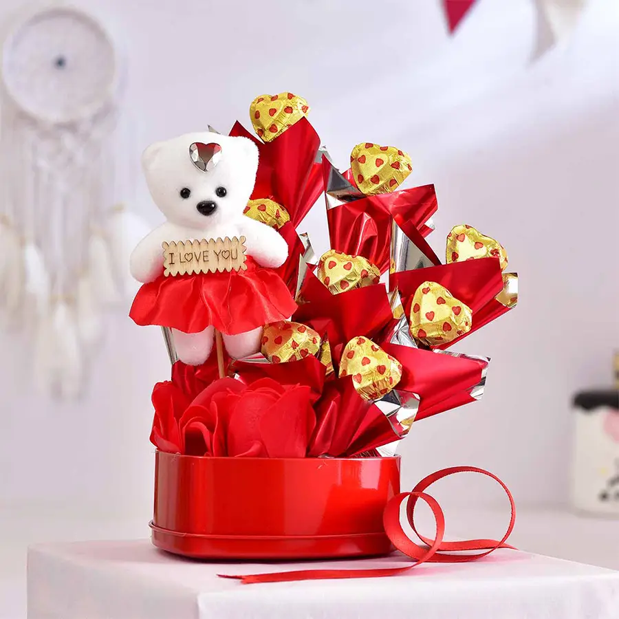 Love Filled Valentine’s Arrangement featuring a red heart-shaped tin with homemade chocolates, a plush doll, fresh roses, and a Valentine’s Day card.