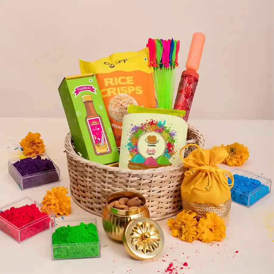 Holi Joy Basket Bonanza filled with festive treats including Phool Thandai, Bluff rice crisps, Chakli, a vibrant Pichkari, water balloons, Gulal in a yellow potli, and 100gms of almonds in a handi jar, all arranged in a rustic cane basket.