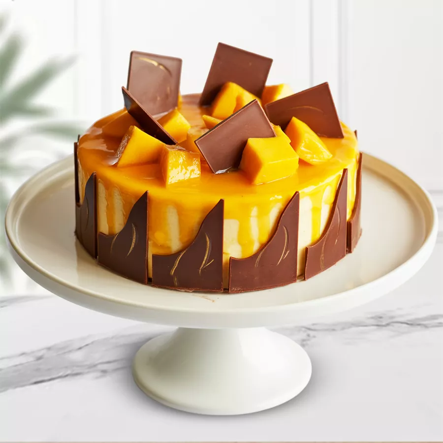 Round eggless mango cake with chocolate base, mango syrup, and chocolate designs, topped with chocolate thins and mango chunks.