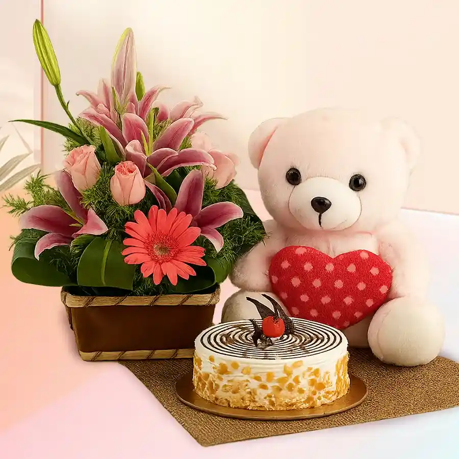Gift basket featuring pink flowers, a butterscotch cake, and a soft teddy bear, perfect for special occasions.