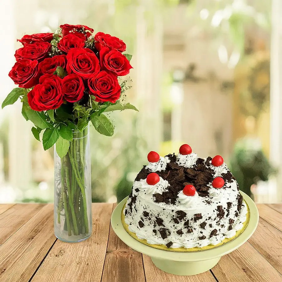A beautiful arrangement of 12 red roses in a vase alongside a round Black Forest cake.