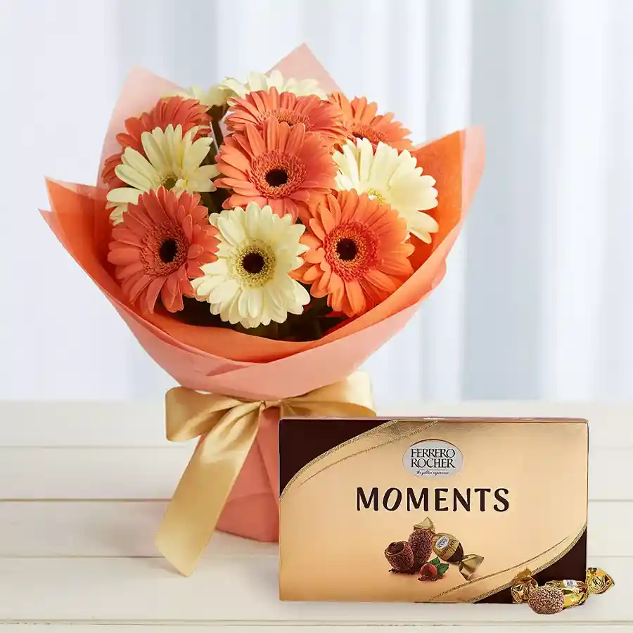 A stunning arrangement of reddish pink and white gerberas in a glass vase with a yellow ribbon, accompanied by a box of Ferrero Rocher chocolates.