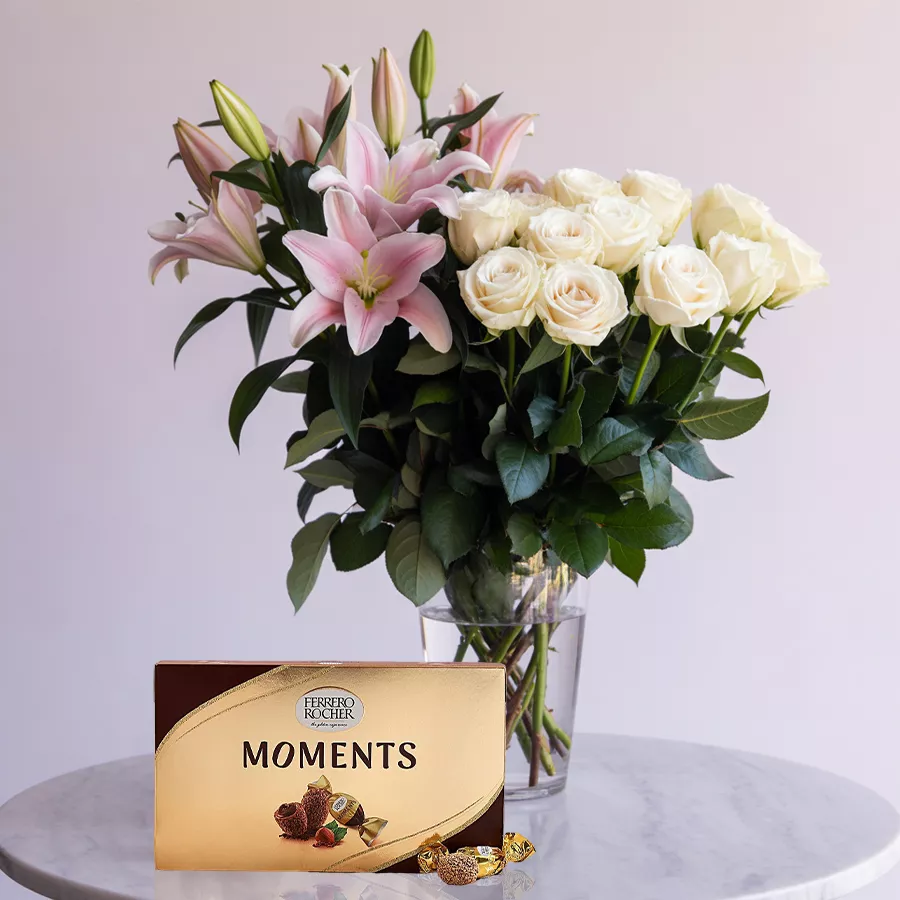 Elegant arrangement of pink lilies and white roses in a glass vase, accompanied by Ferrero Rocher chocolates.