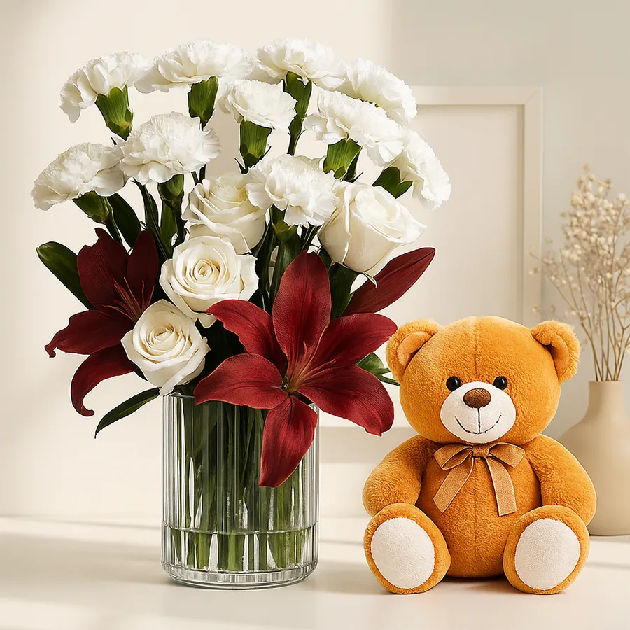 Artistic flowers display with white carnations, roses, red anthuriums, and a cute teddy bear in a glass vase.