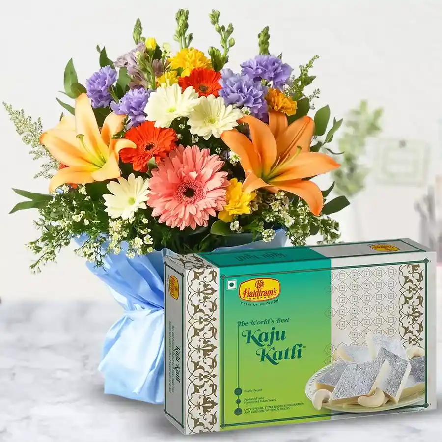 A delightful gift set featuring Kaju Barfi and a bouquet of seasonal flowers including Lilies, Gerberas, and Carnations.