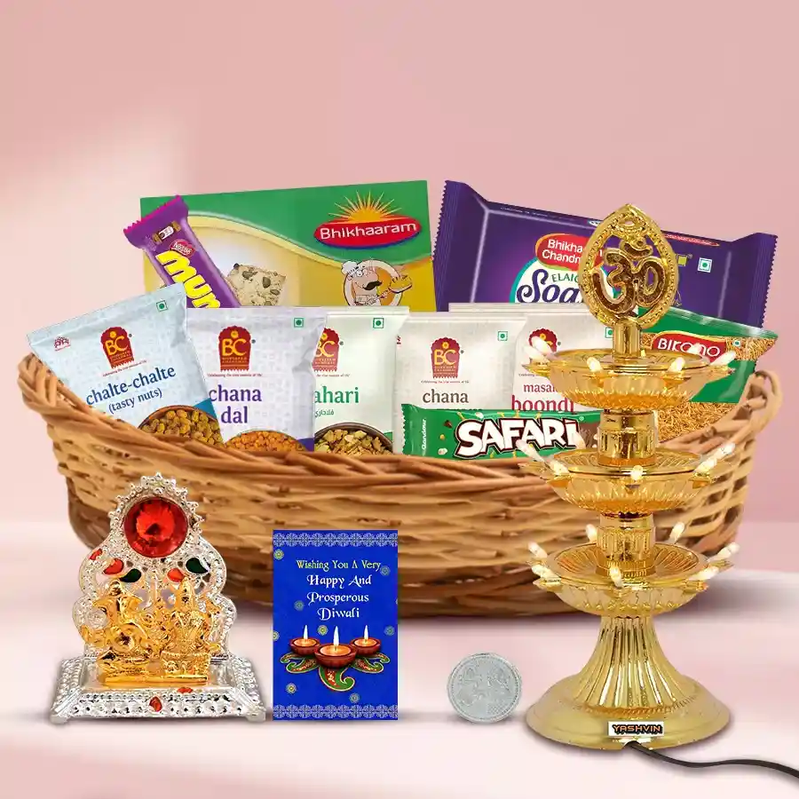 A beautifully arranged Diwali gift basket featuring Bhikaram Namkeen, Soan Papdi, baked Kaju Pista bites, chocolate bars, a Ganesh-Laxmi Mandap, a 3-tier LED lamp, a silver-plated Ganesh-Laxmi coin, all presented in a reusable cane basket with a greetings card.