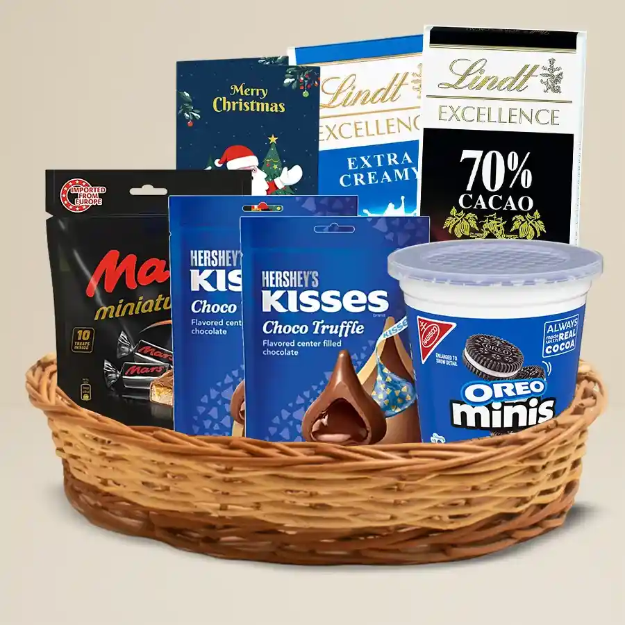 A beautifully arranged gift basket filled with assorted chocolates including Hershey’s, Cadbury, Mars, and Lindt, accompanied by a Christmas greeting card, all presented in a stylish cane basket.