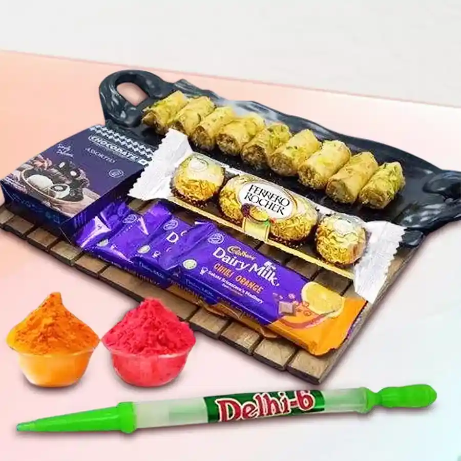 A colorful gift bundle featuring Baklawa sweets, Cadbury Dairy Milk Chilli Orange bars, Ferrero Rocher chocolates, a chocolate box, a long pitchkari, and herbal gulal, all arranged festively for Holi celebrations.