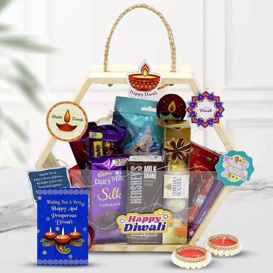 A beautifully arranged gift hamper featuring an assortment of chocolates including Hershey’s Kisses, Ferrero Rocher, Cadbury chocolates, and Nestle Kit Kats, alongside a silver Ganesh Laxmi coin, elegant candle holders, and a hexagonal basket, with a free Diwali card included.