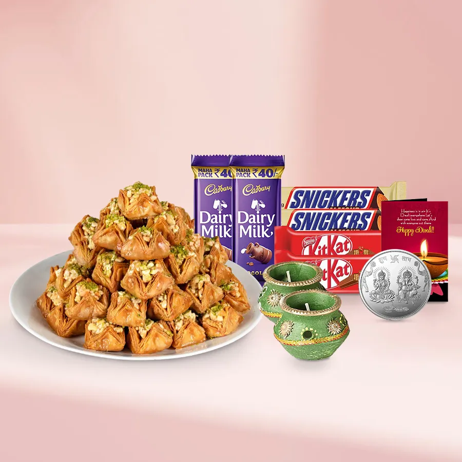A decorative basket filled with Pyramid Baklava, Snickers, Cadbury Dairy Milk, KitKat bars, festive wax diyas, and a silver Ganesh Laxmi coin, topped with a Diwali card.