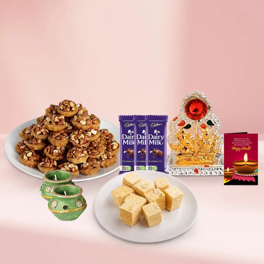 A beautifully arranged decorative basket containing Tart Baklava, Soan Papdi, Cadbury Dairy Milk chocolates, decorated wax diyas, and a Ganesh Lakshmi Mandap, alongside a Diwali card.