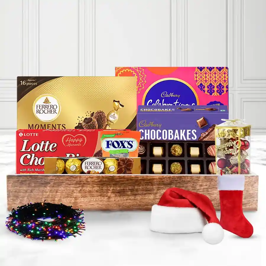 A festive gift tray filled with Lotte Chocopie, Cadbury Celebration, Ferrero Rocher, handmade chocolates, Christmas decorations, twinkling lights, and fun caps and stockings, all arranged on a reusable wooden tray.