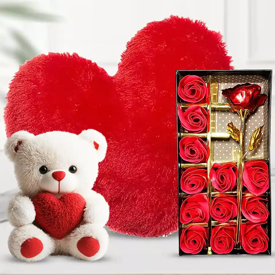 Teddy bear holding a heart, red roses in an elegant box, and a heart-shaped cushion arranged together as a gift set.