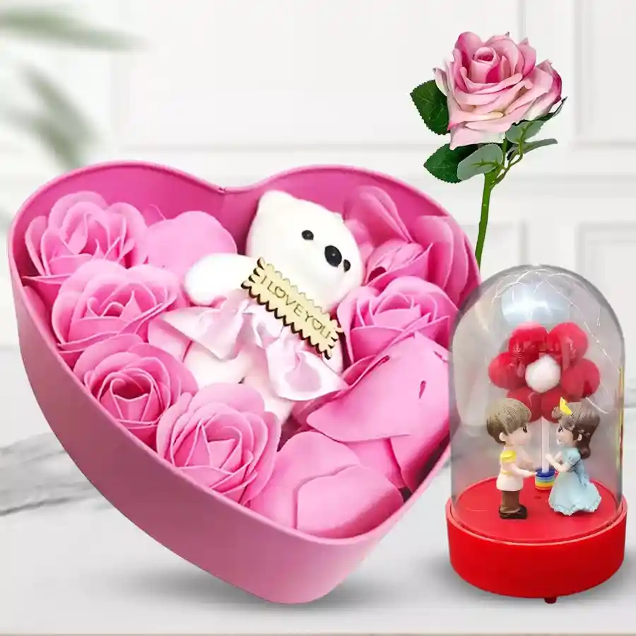 Heart-shaped box filled with vibrant roses, a soft teddy bear, a couple showpiece in a glowing glass bottle with LED light, and a single pink rose on a stick.