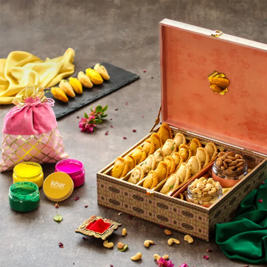 A pink floral box filled with traditional Holi sweets, including gujiya and nuts, alongside vibrant herbal colors and a greeting card.