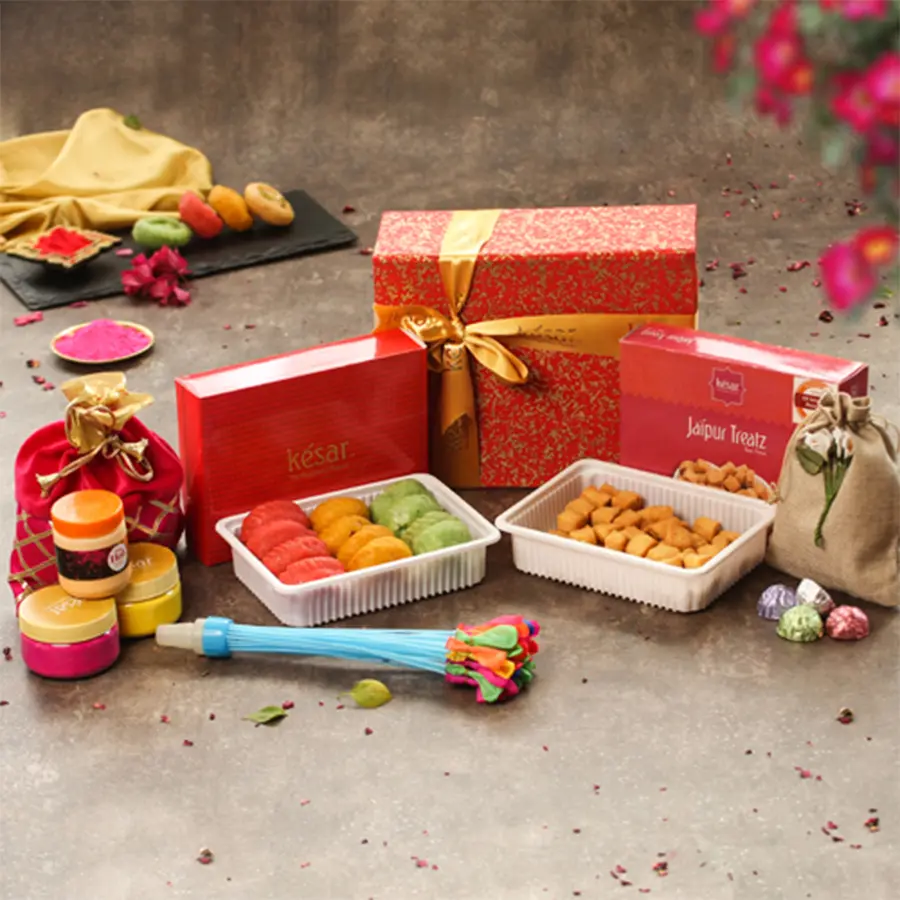 Tasty assorted sweets and chocolates in a handmade paper box with herbal colors, pichkari balti, water balloons, and a greeting card for Holi celebration.
