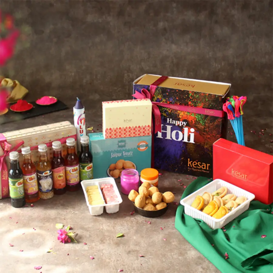 Flavorsome Treat Gift Hamper for Holi featuring assorted gujiyas, kachoris, thandai, kaju katli, gulal, chandan, water balloons, color bomb, and a greeting card in a colorful box.
