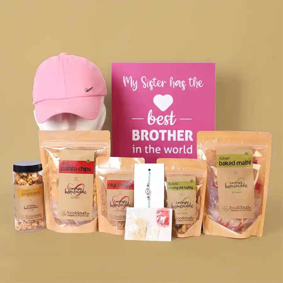 Gift box containing snacks like potato chips, baked mathi, moong dal twists, ragi bhujiya, and caramel popcorn alongside a pink cap, a designer Evil Eye Rakhi, and roli chawal for Raksha Bandhan celebration.