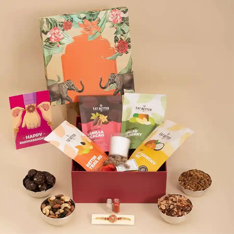 A beautifully arranged gift hamper for Raksha Bandhan featuring a handcrafted Rakhi, gluten-free treats including chocolate laddoos and trail mixes, a soy wax candle, seed bomb, and decorative glass bottles with Roli, Tilak, and Chawal, all complemented by a heartfelt greeting card.