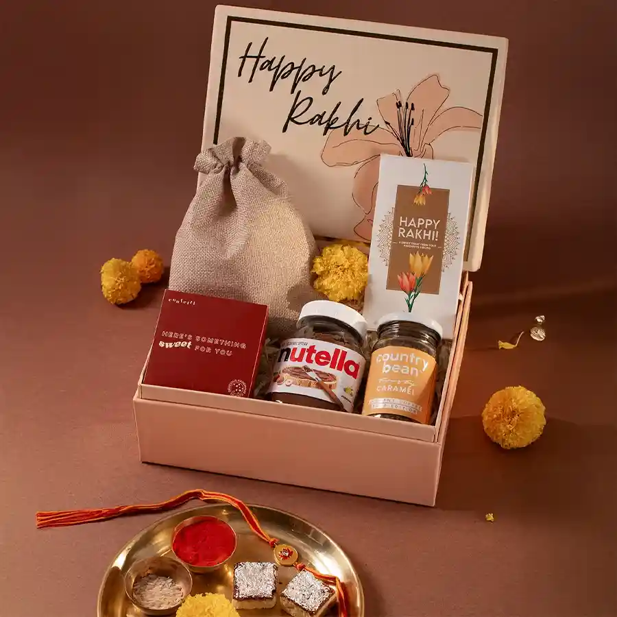 Gourmet box containing caramel flavored coffee, premium dry fruits, handmade choco-chip cookies, Nutella spread, Happy Rakhi chocolate bar, Kundan Rakhi, and a peach box, styled for Raksha Bandhan celebration.