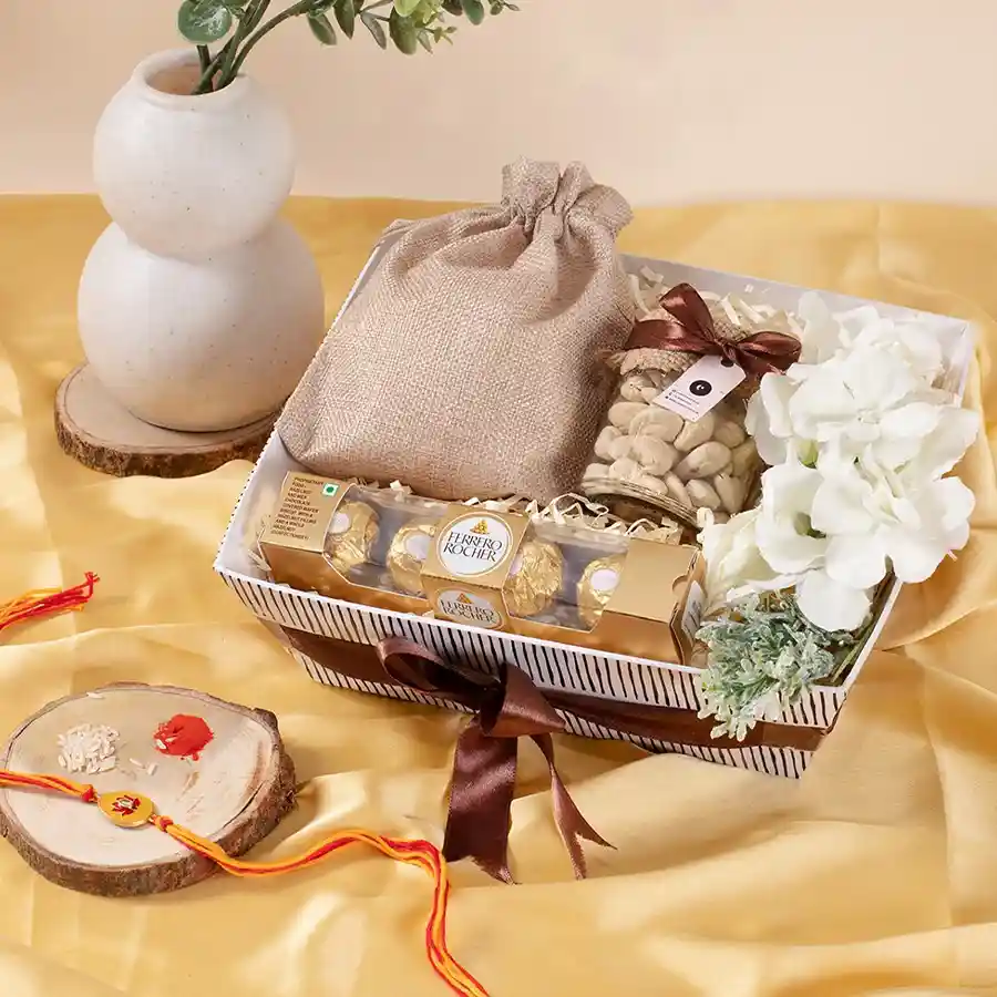 Nuts n Chocolates Rakhi Gift featuring cashews in a glass jar, seed mix in a jute potli, Ferrero Rocher chocolates, a flower arrangement, reusable kappy tray, and a meenakari rakhi.