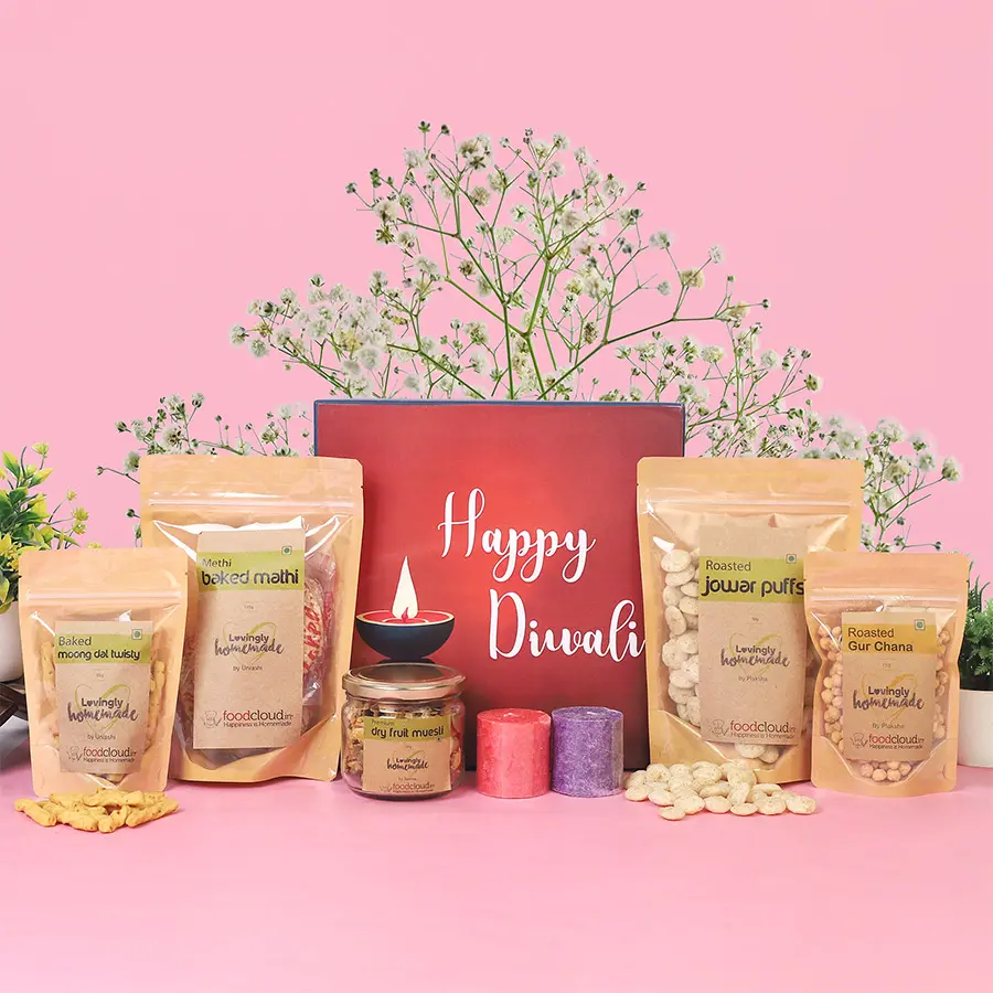 A beautifully arranged Festive Delights Diwali Celebration Box featuring various snacks like Roasted Gur Chana, Dry Fruit Muesli, Baked Moong Dal Twisty, Roasted Jowar Puffs, Baked Mathi, and two scented candles.