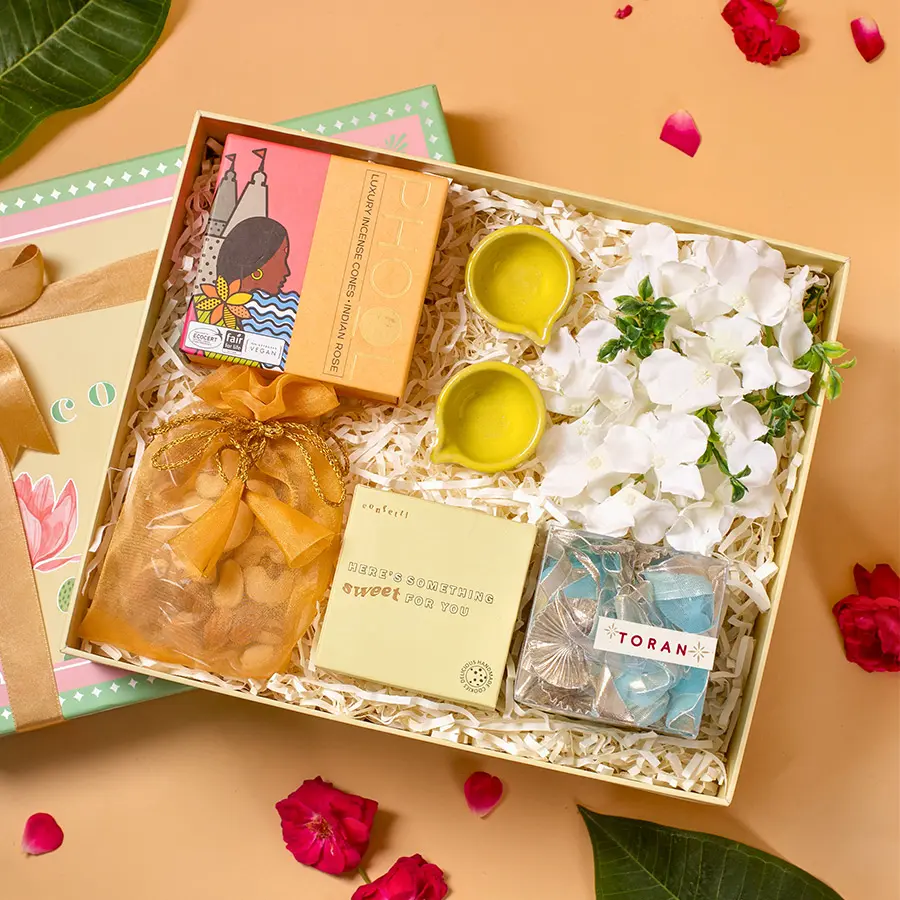 A beautifully arranged gift box for Diwali featuring a blue toran, phool incense cones, green ceramic diyas, handmade nut cookies, premium dry fruits in an organza potli, vibrant flower decor, and a chic green lotus box.