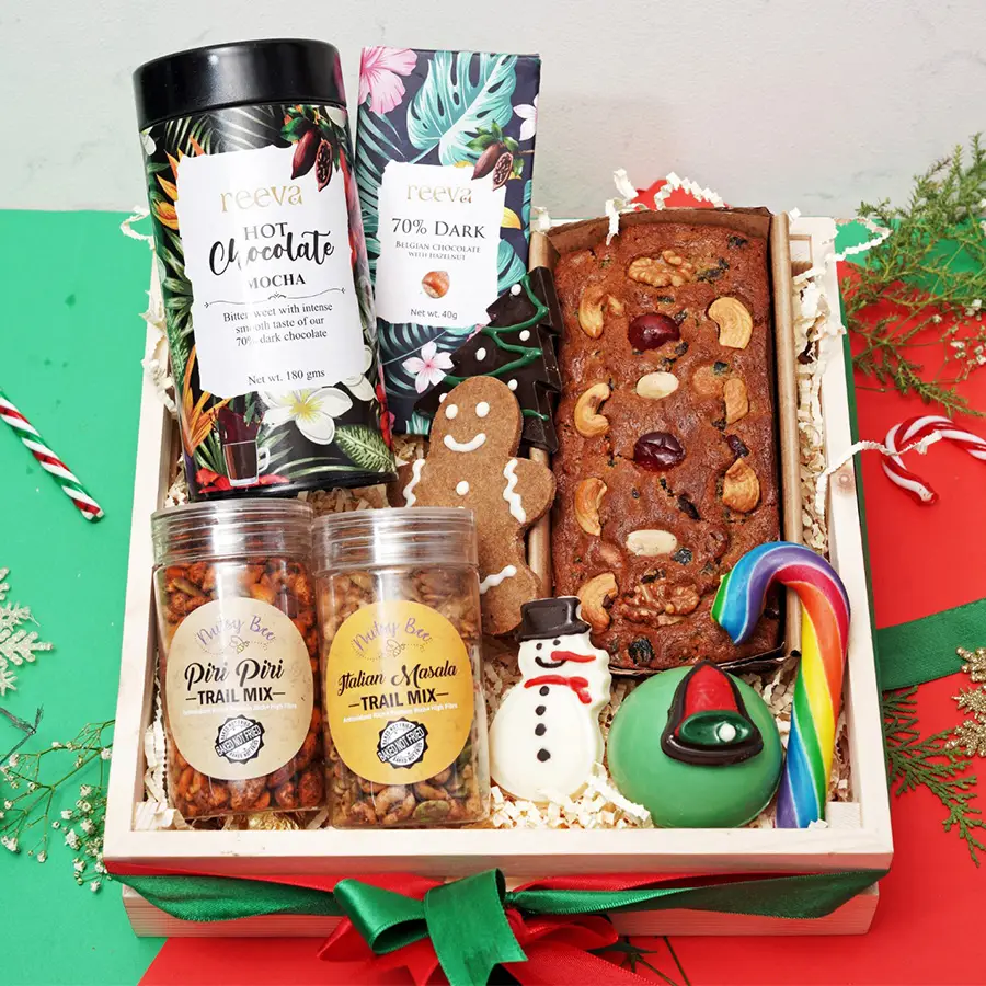 Christmas Magic Treats Gift Set featuring hot chocolate, dark chocolate bar, gingerbread cookie, fruit and nut tea cake, trail mix, snowman figure, hot chocolate bomb, and candy cane arranged in a festive display.