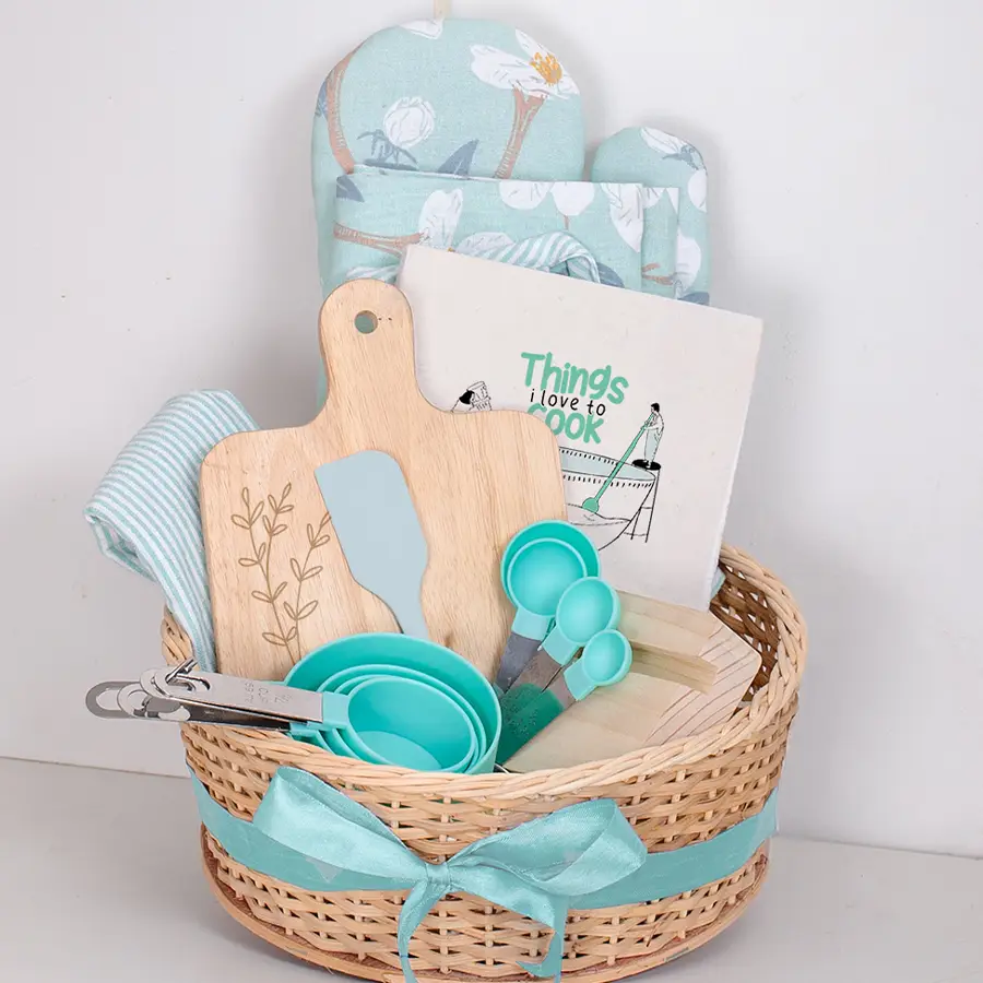 Set of kitchen essentials including a wooden phone stand, cooking book, spatula, measuring cups and spoons, rubber wood chopping board, chic apron set, and reusable cane basket, arranged in a decorative gift box.
