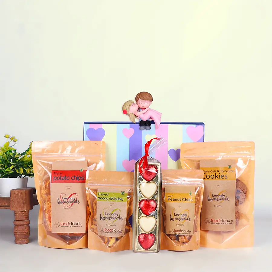 Joyful Munchies Gift Hamper featuring Couple Miniatures, Gur Peanut Chikki, Oats & Choco Chip Cookies, Fiery Potato Chips, Baked Moong Dal Twisty, and heart-shaped white chocolates.