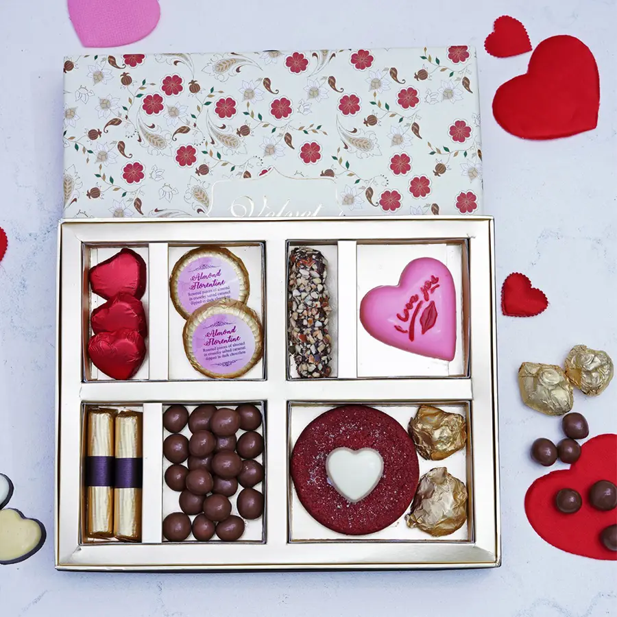 A beautifully arranged gift box filled with assorted chocolates including heart-shaped chocolates, almond chocolate florantines, a chocolate finger, strawberry logs, hazelnut dragees, a red velvet cookie, and chocolate almond rocks.